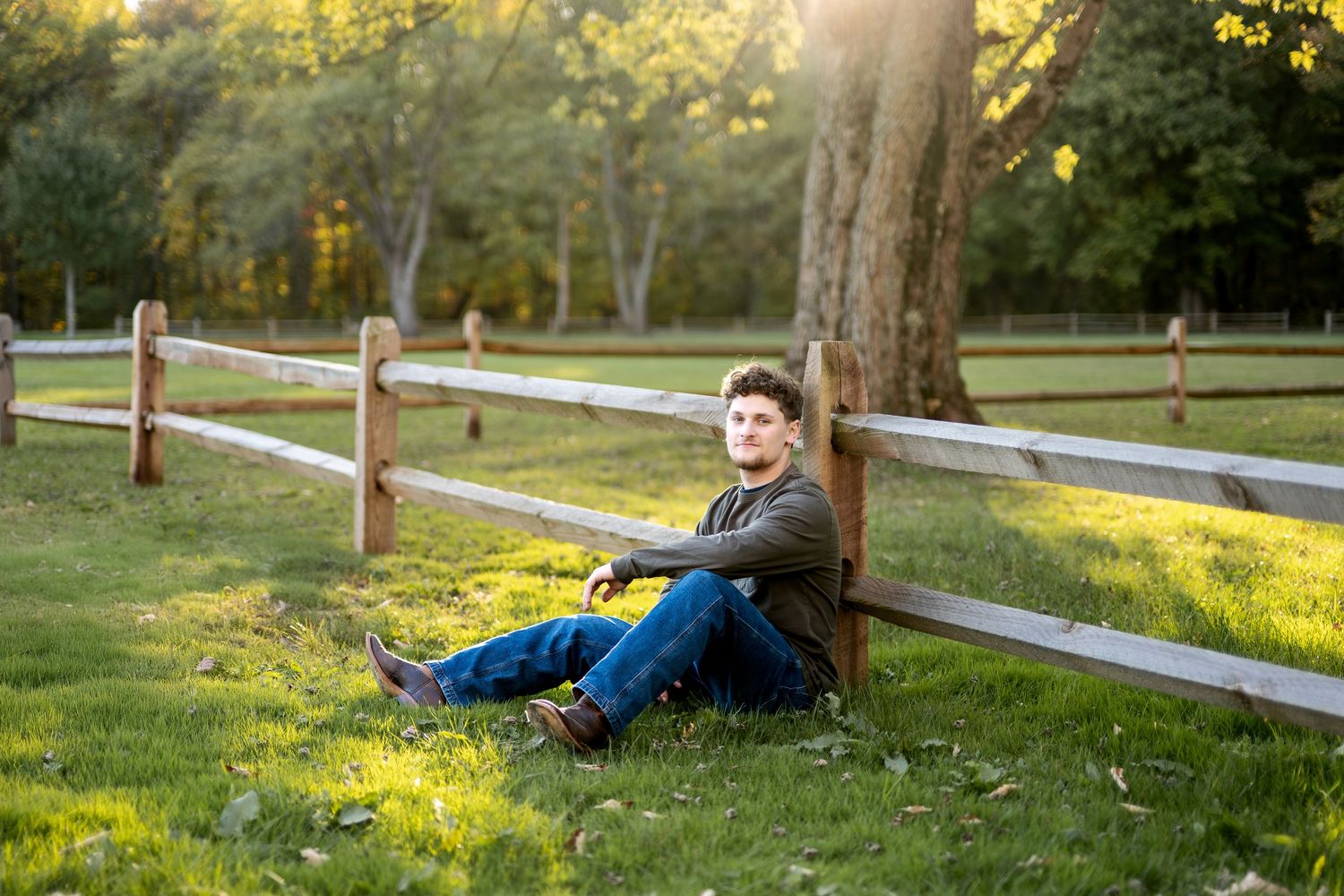 Young man in casual clothes sits relaxed against a wooden fence post on a sunny green lawn.