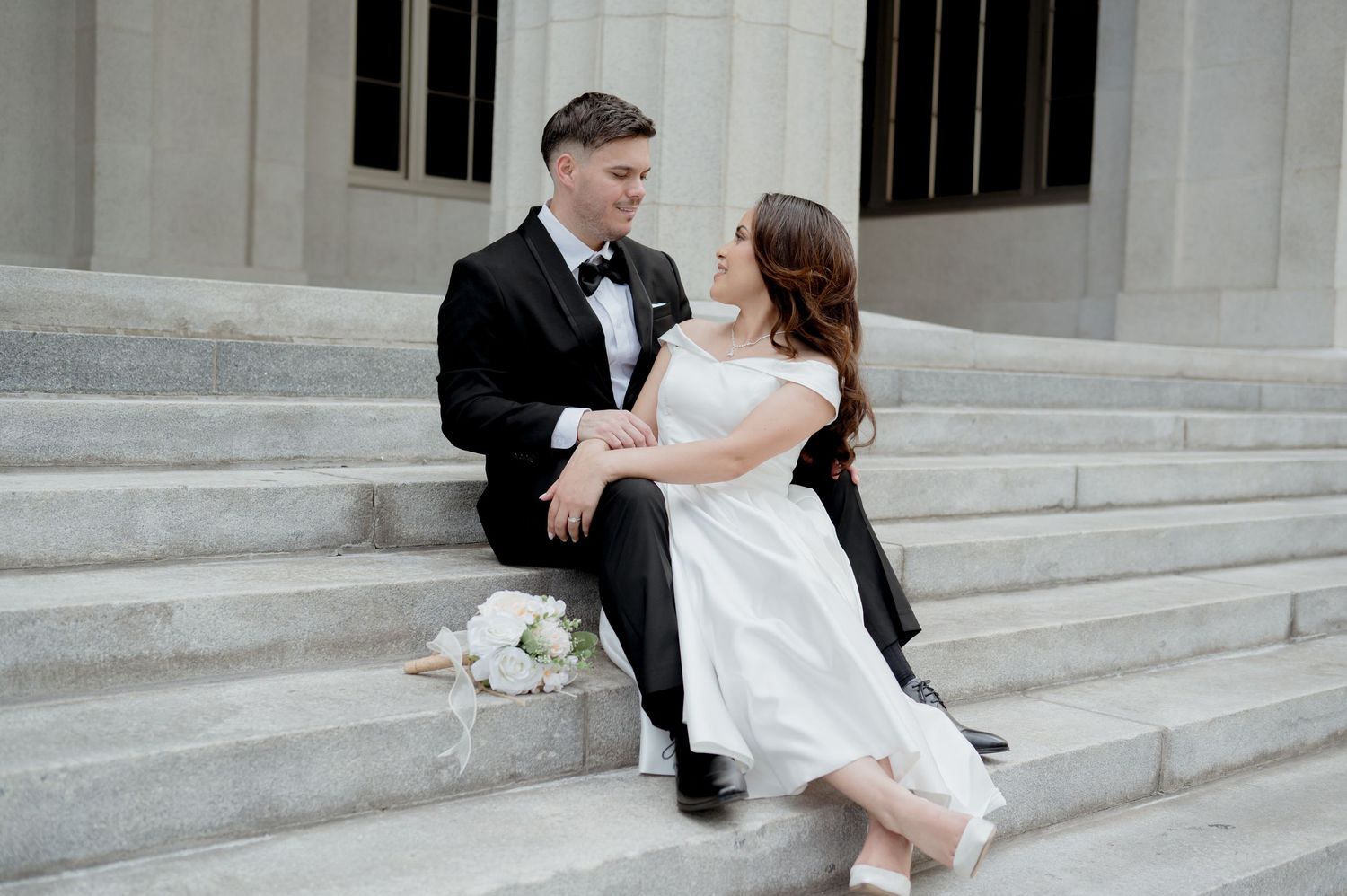 Newlywed couple sits romantically on stone steps with bridal bouquet, bride in white dress and groom in classic tuxedo.