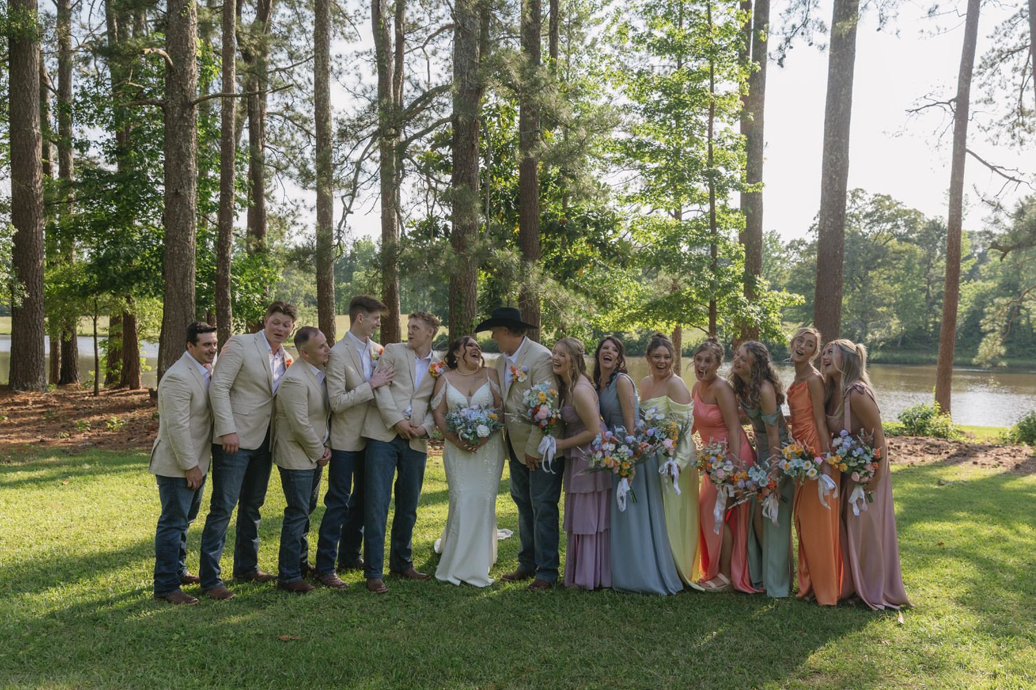 Wedding party in pastel dresses and khaki suits poses together on a sunny lawn near a lake.