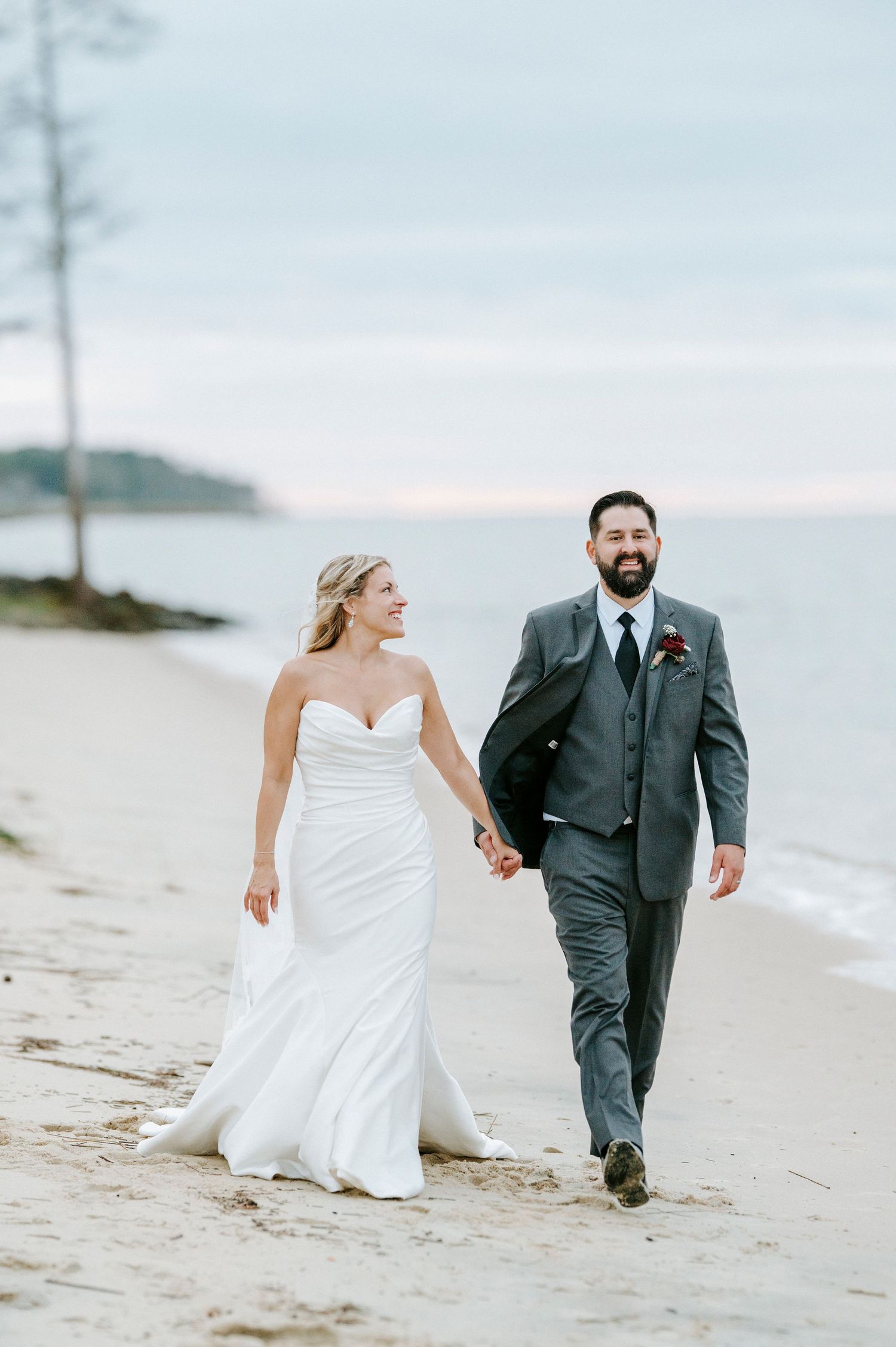 A couple walks hand in hand along a beach at sunset, with the bride in white and groom in charcoal gray suit.