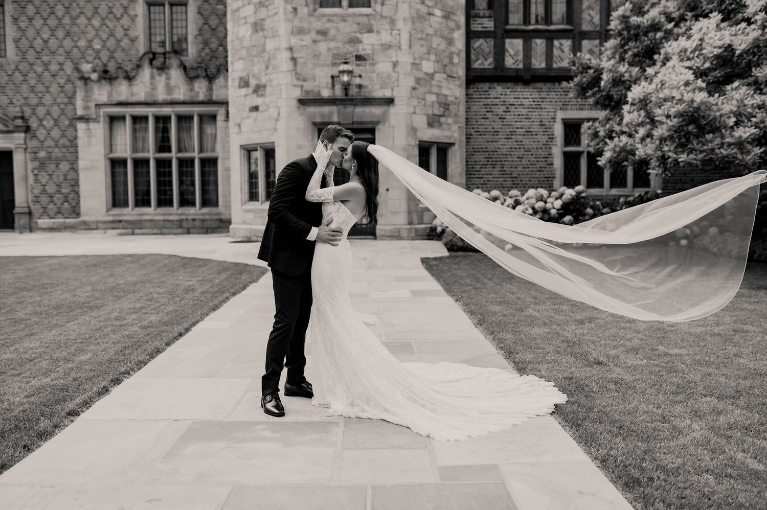 Bride and groom share a romantic kiss outdoors at a grand estate, with the veil flowing dramatically in the wind.
