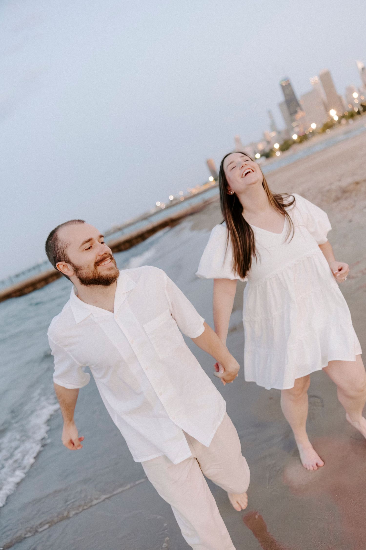 A couple in white clothing laughs and plays together on a beach at sunset with twinkling lights in the background.