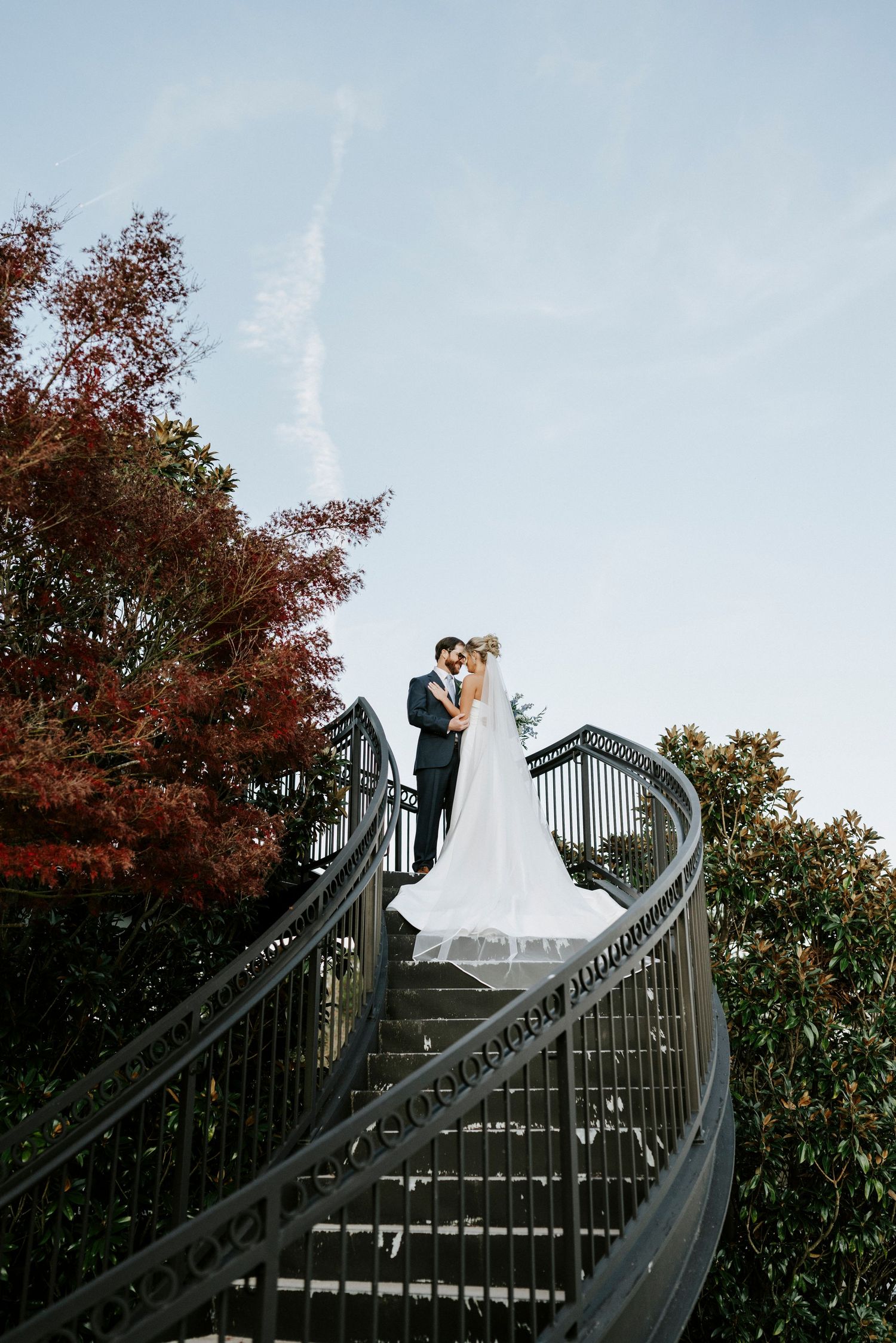 A couple embraces at the top of an ornate metal staircase surrounded by autumn foliage at sunset.