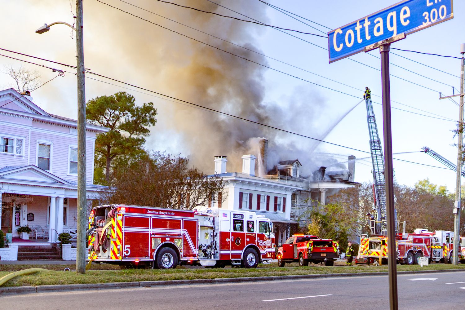 Multiple fire trucks and emergency vehicles respond to a house fire with smoke billowing from residential buildings.