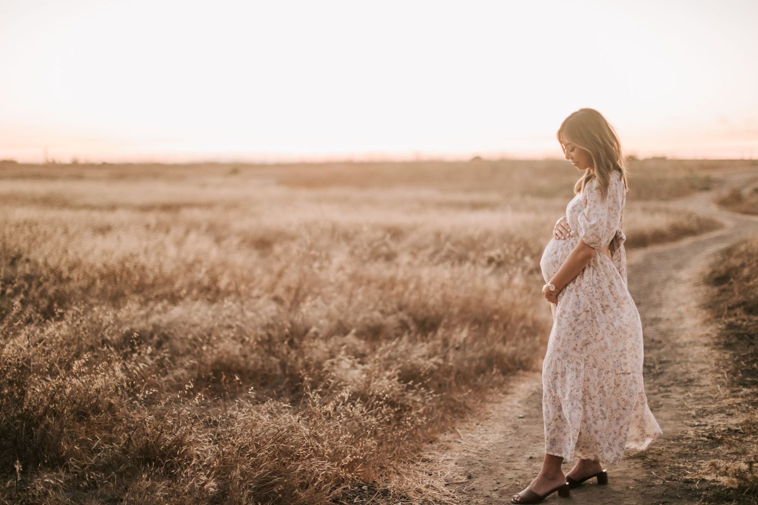 Woman in flowy white dress walks along rocky beach shoreline during golden sunset photography session.