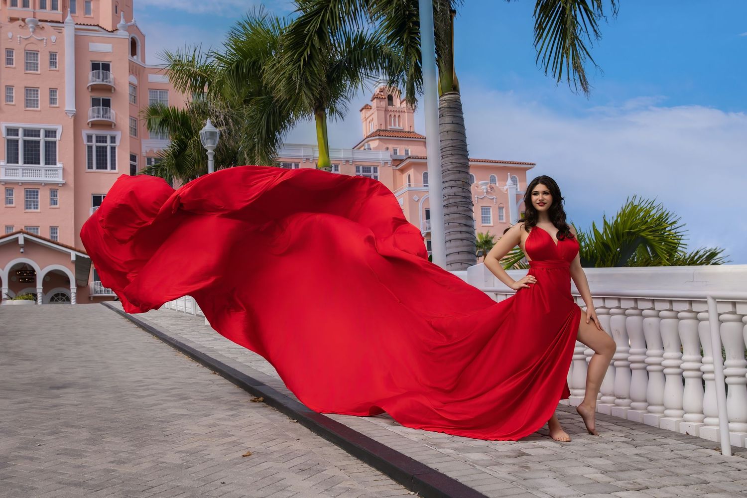 A person in a flowing red dress poses dramatically against an ornate building exterior with palm trees.