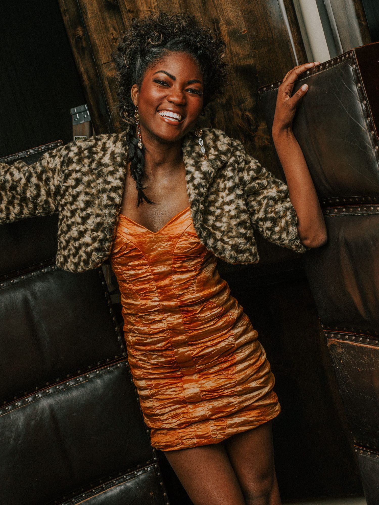 A stylish portrait featuring a vibrant orange ruched dress paired with a leopard print jacket against a dark background.