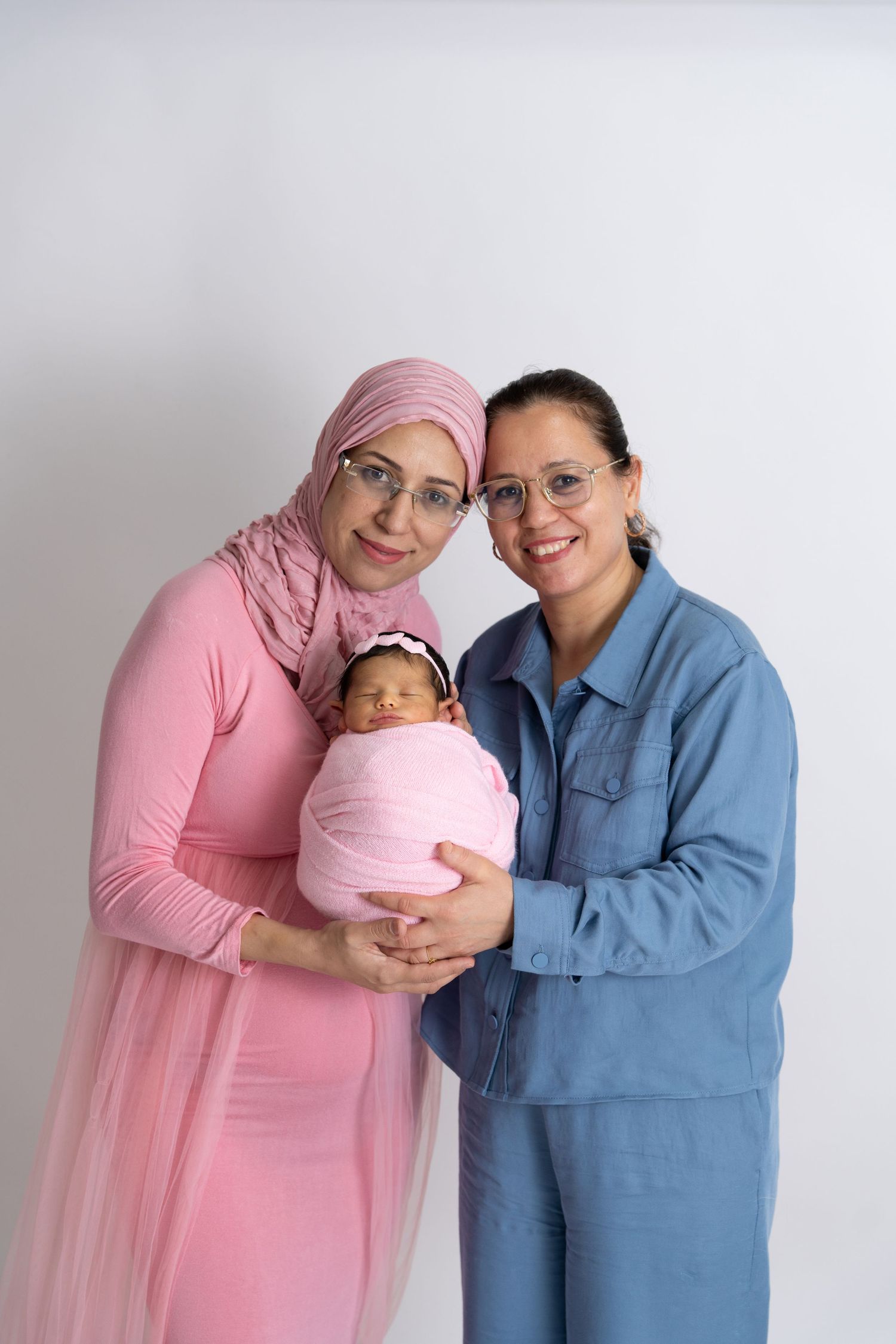 A newborn baby wrapped in pink blanket being held by two adults wearing pink and blue outfits against white background.