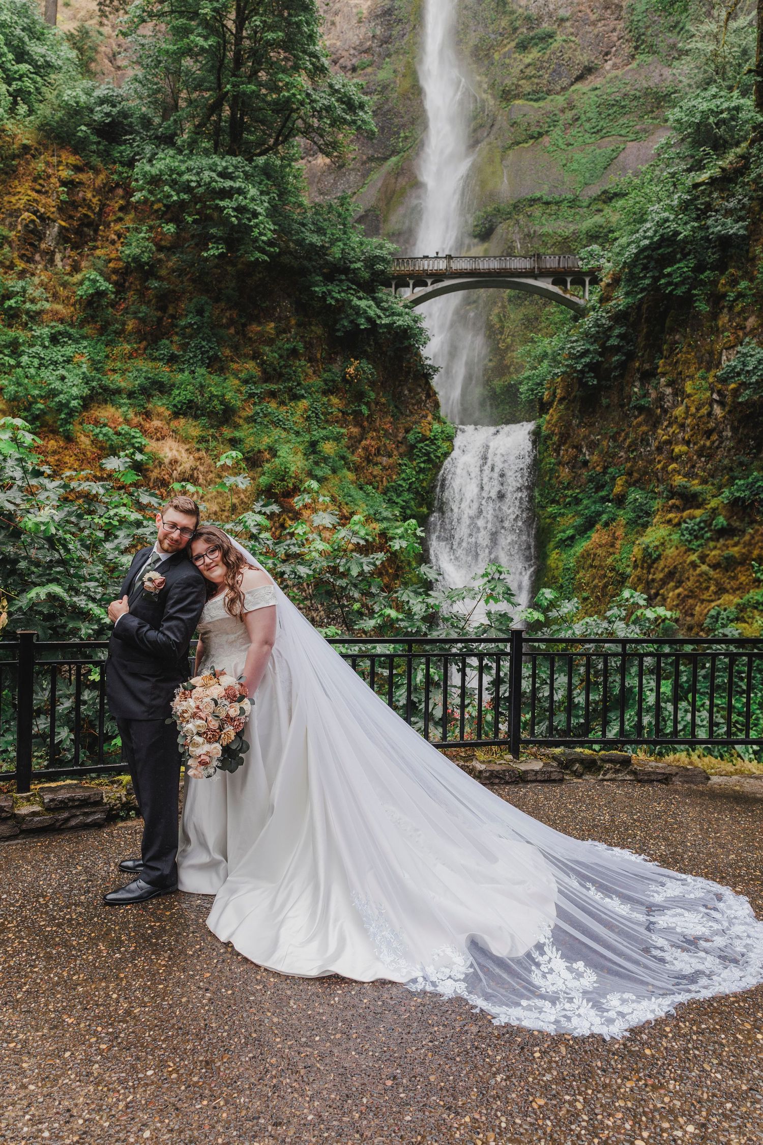 Two people in wedding attire pose at the scenic Multnomah Falls viewing bridge in Oregon.