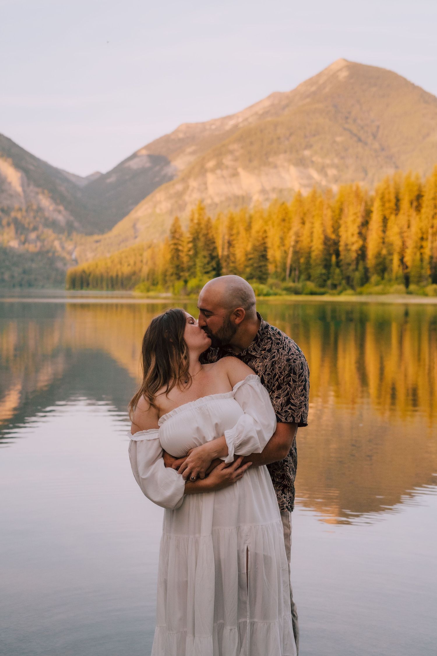 A couple embraces by a serene mountain lake at sunset with golden larches reflected in the still water.
