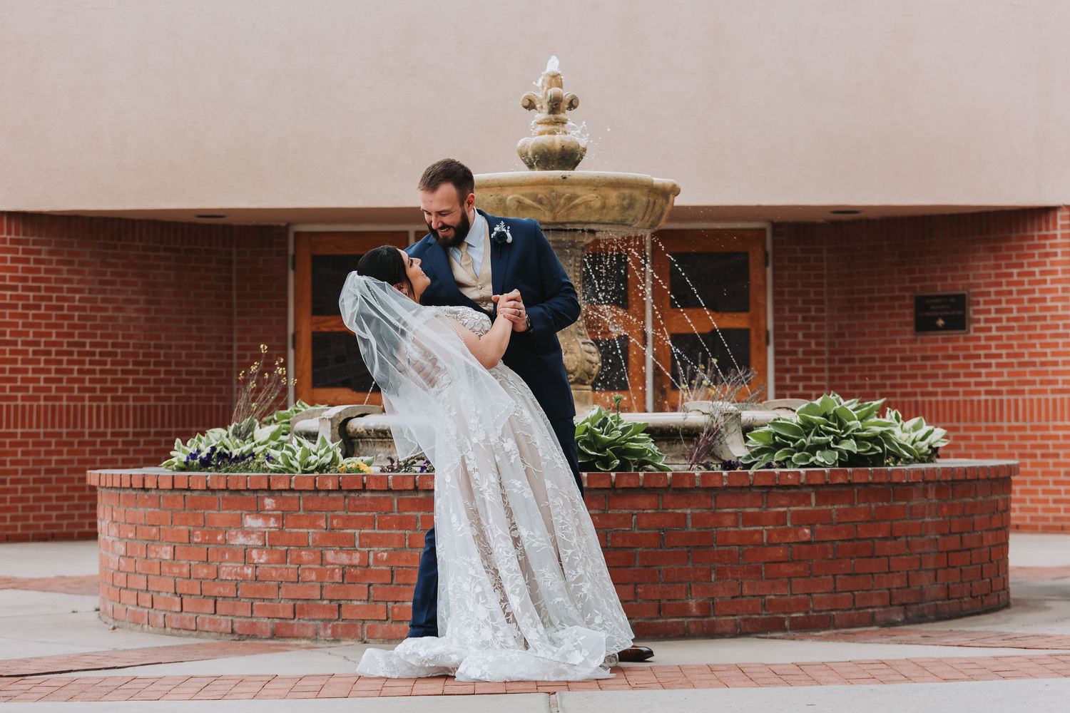 A romantic dip pose in front of a brick fountain with decorative plants and greenery.