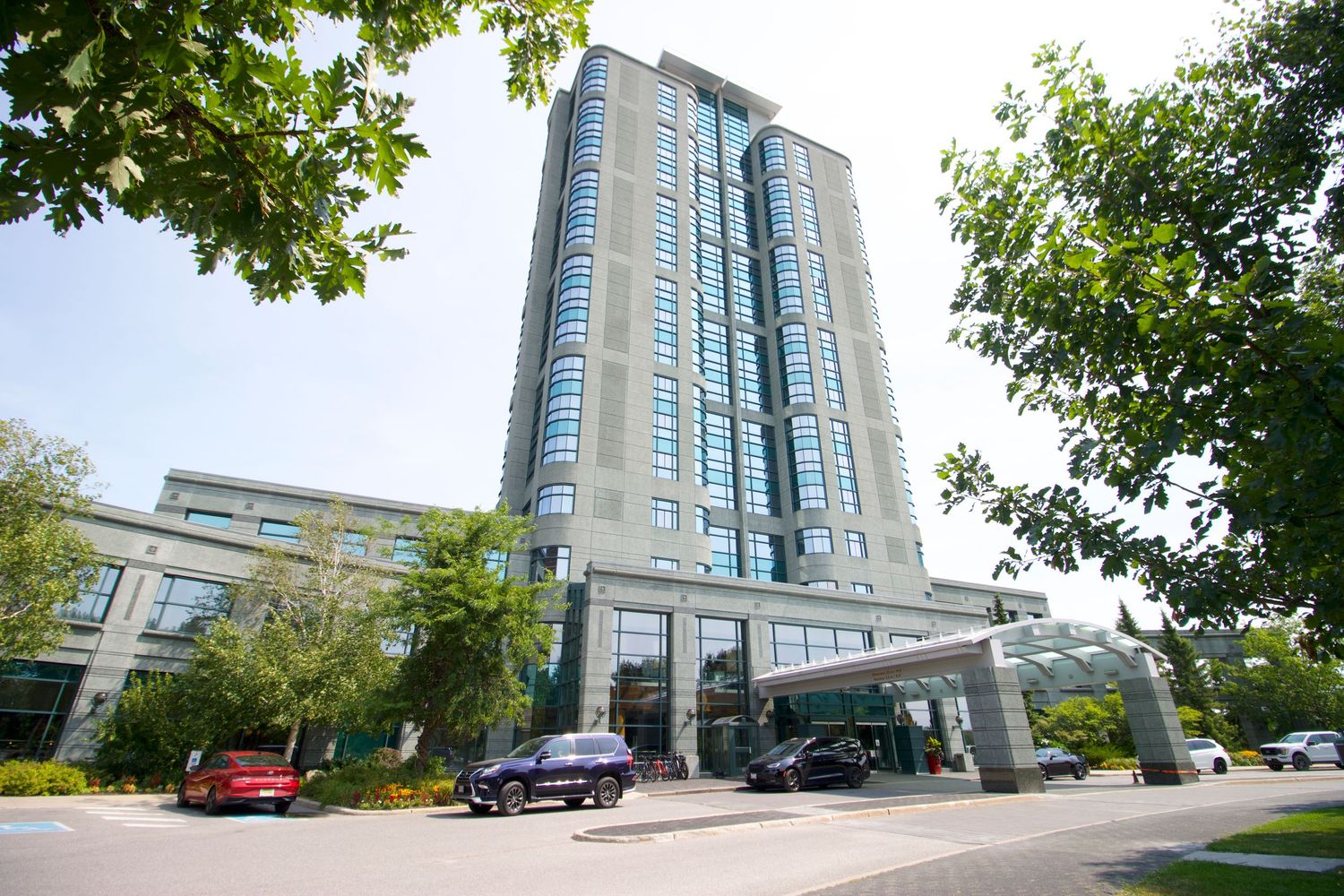 Modern glass high-rise building with blue tinted windows and covered entrance surrounded by trees in an urban setting.