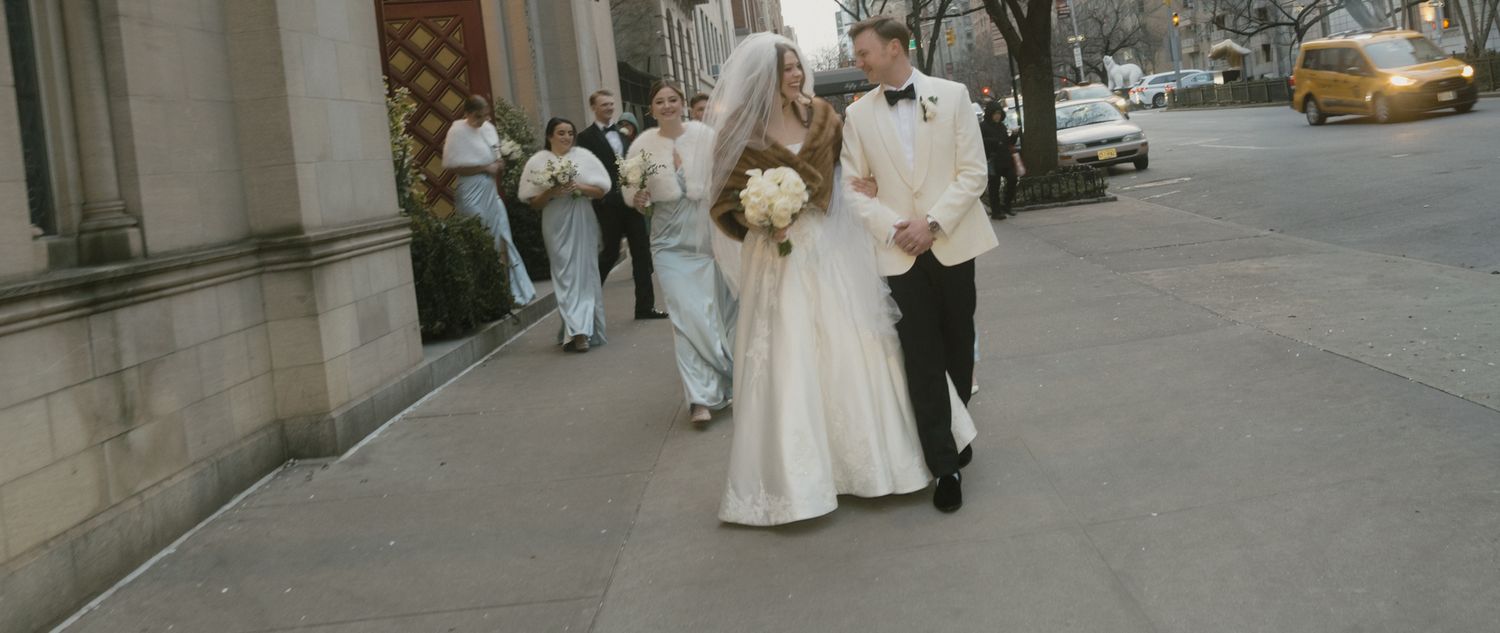 People in wedding attire walk down a city street past stone buildings and a yellow taxi cab.