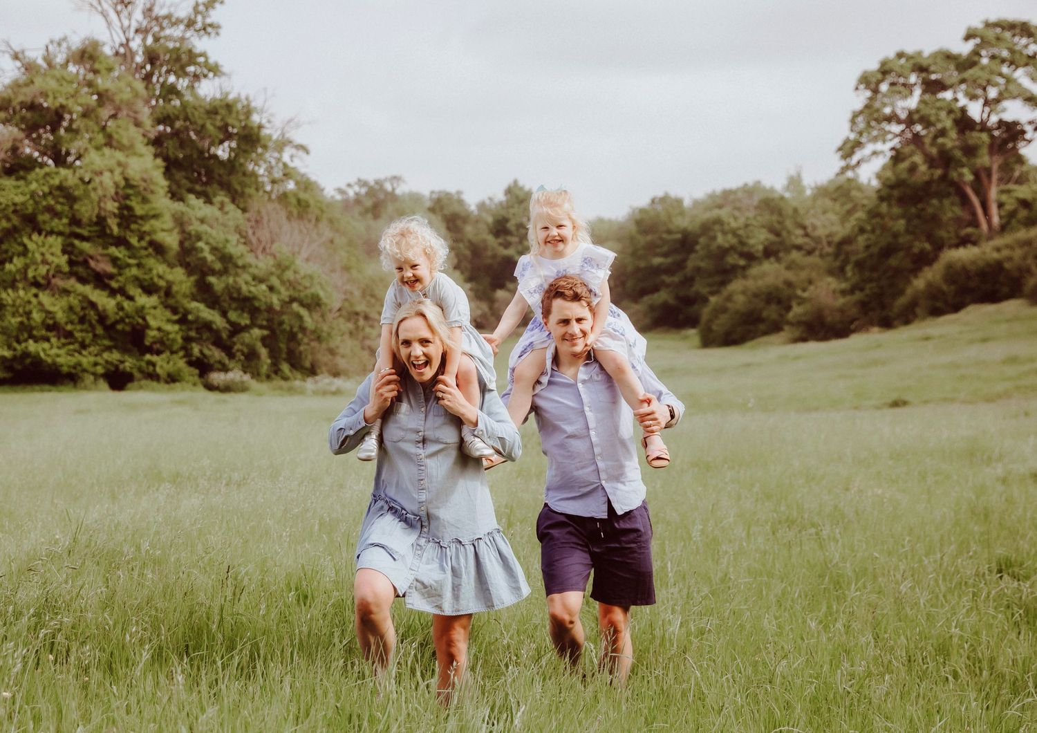 Happy family of four walking in green meadow, parents carrying young daughters on shoulders outdoors during a family photoshoot in London