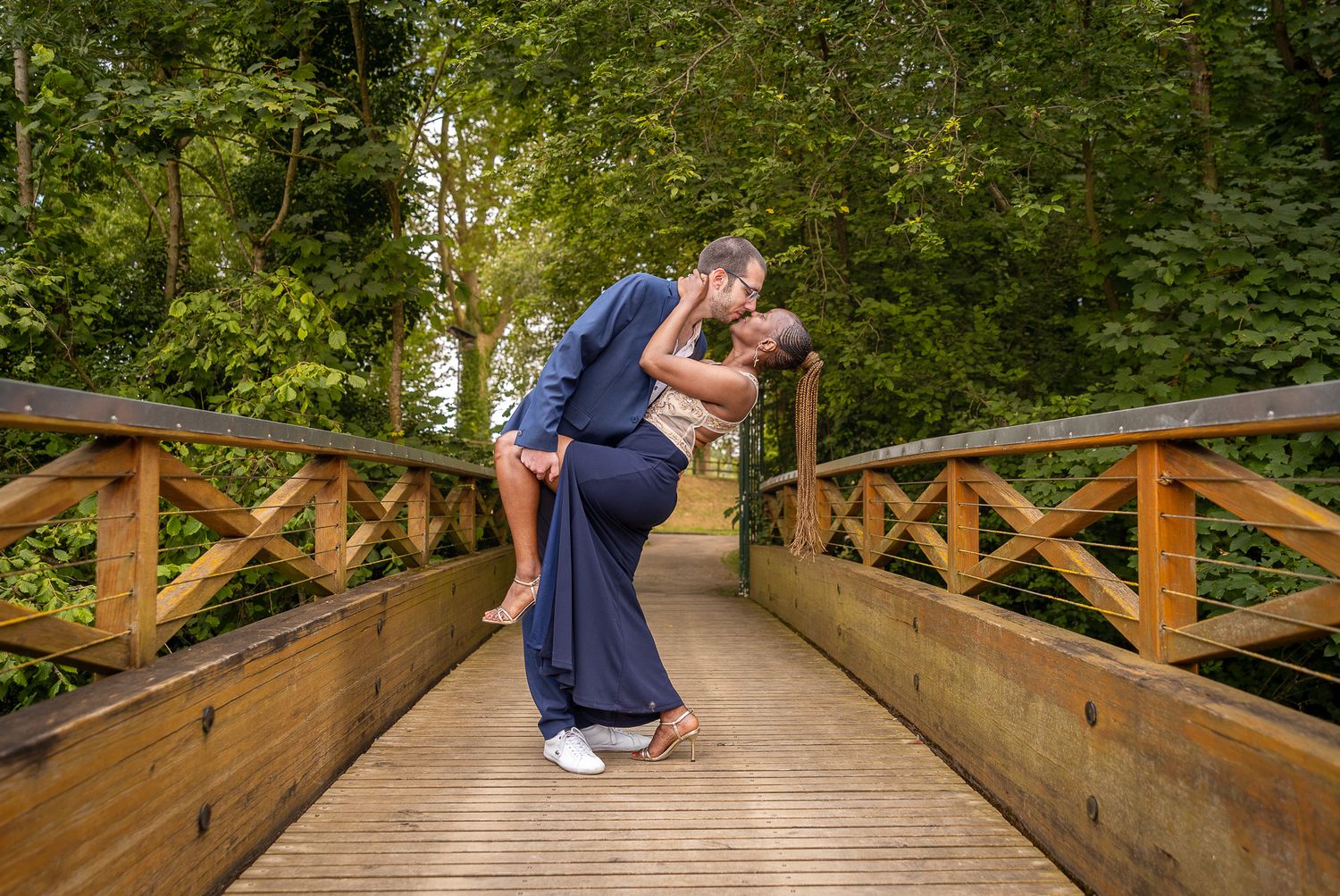 Couple in matching navy outfits sharing a romantic moment while standing on a wooden bridge in a lush forest setting.