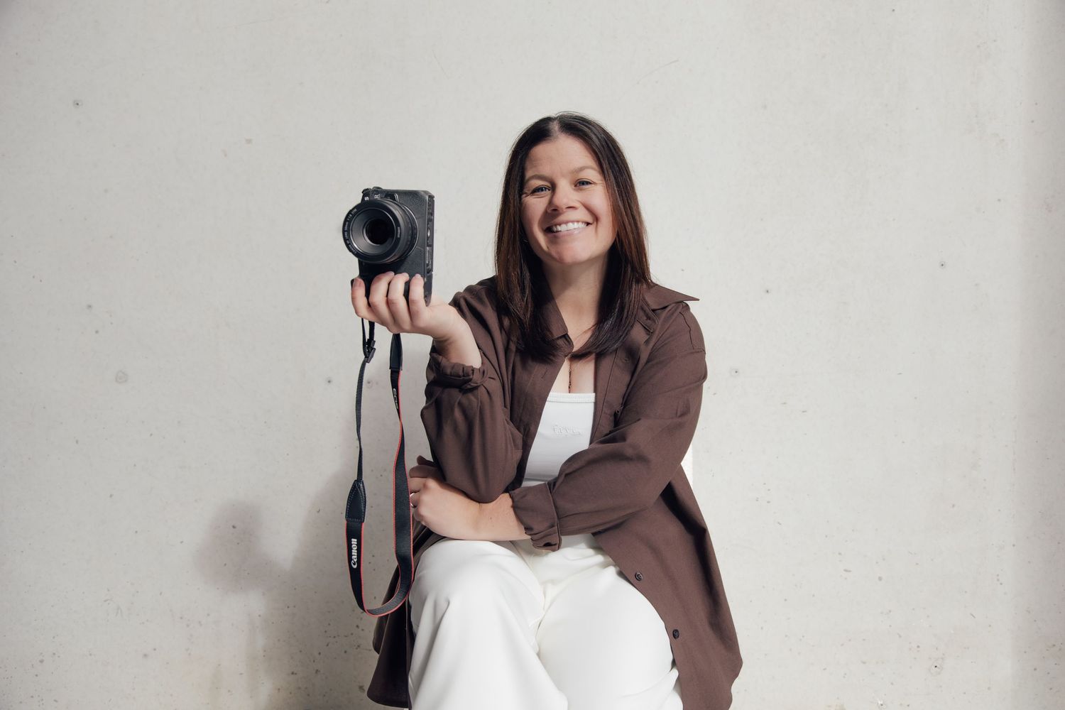 Professional photographer in a brown jacket positions herself in different poses against a white wall with camera.