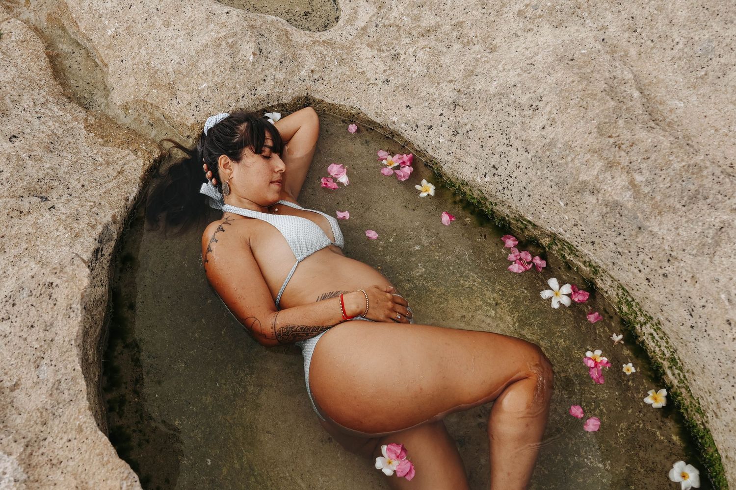 A woman in a white bikini relaxes in a natural rock pool surrounded by tropical flowers and petals.