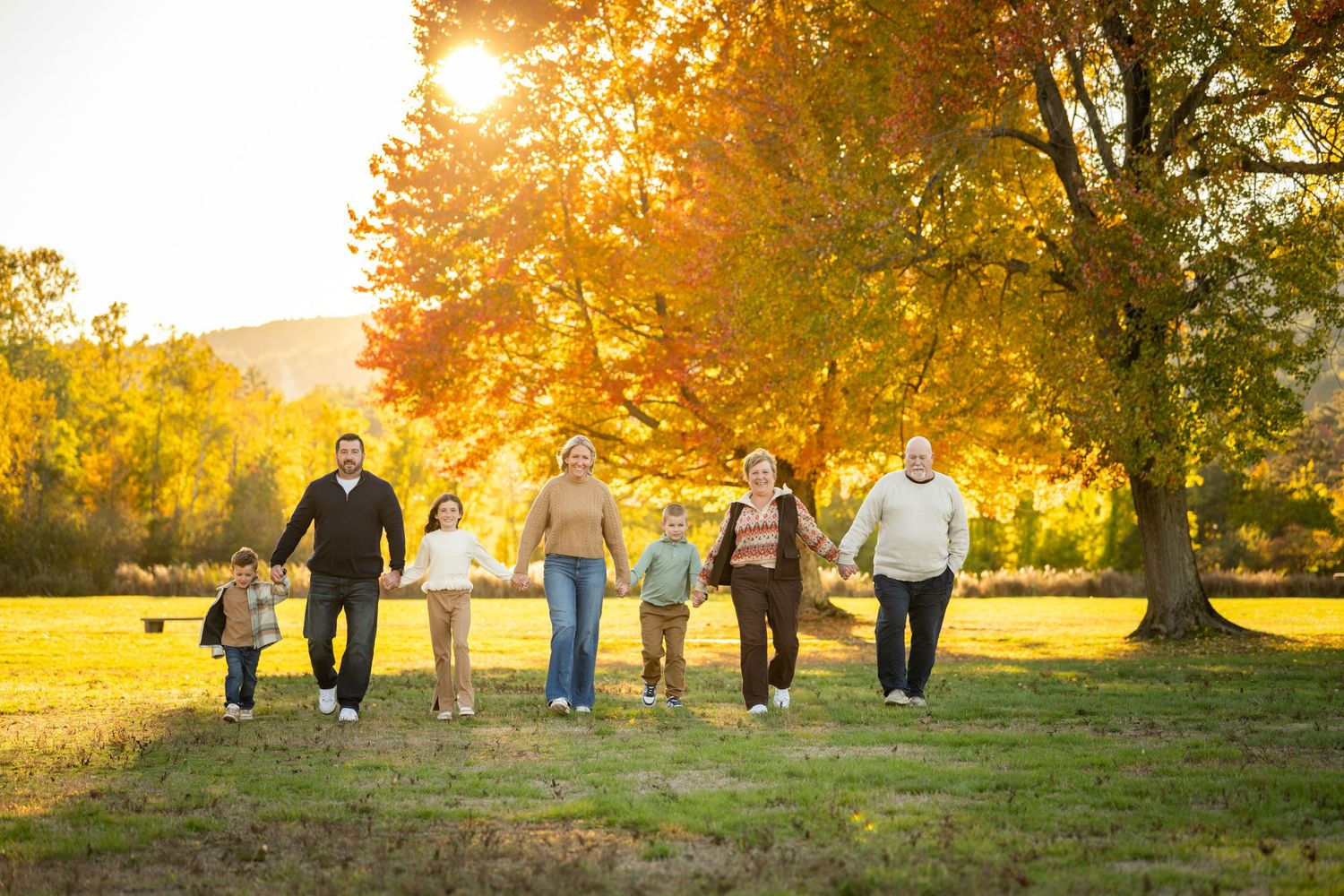 A group walks together holding hands in a park during golden hour with autumn trees and sunlight in the background.