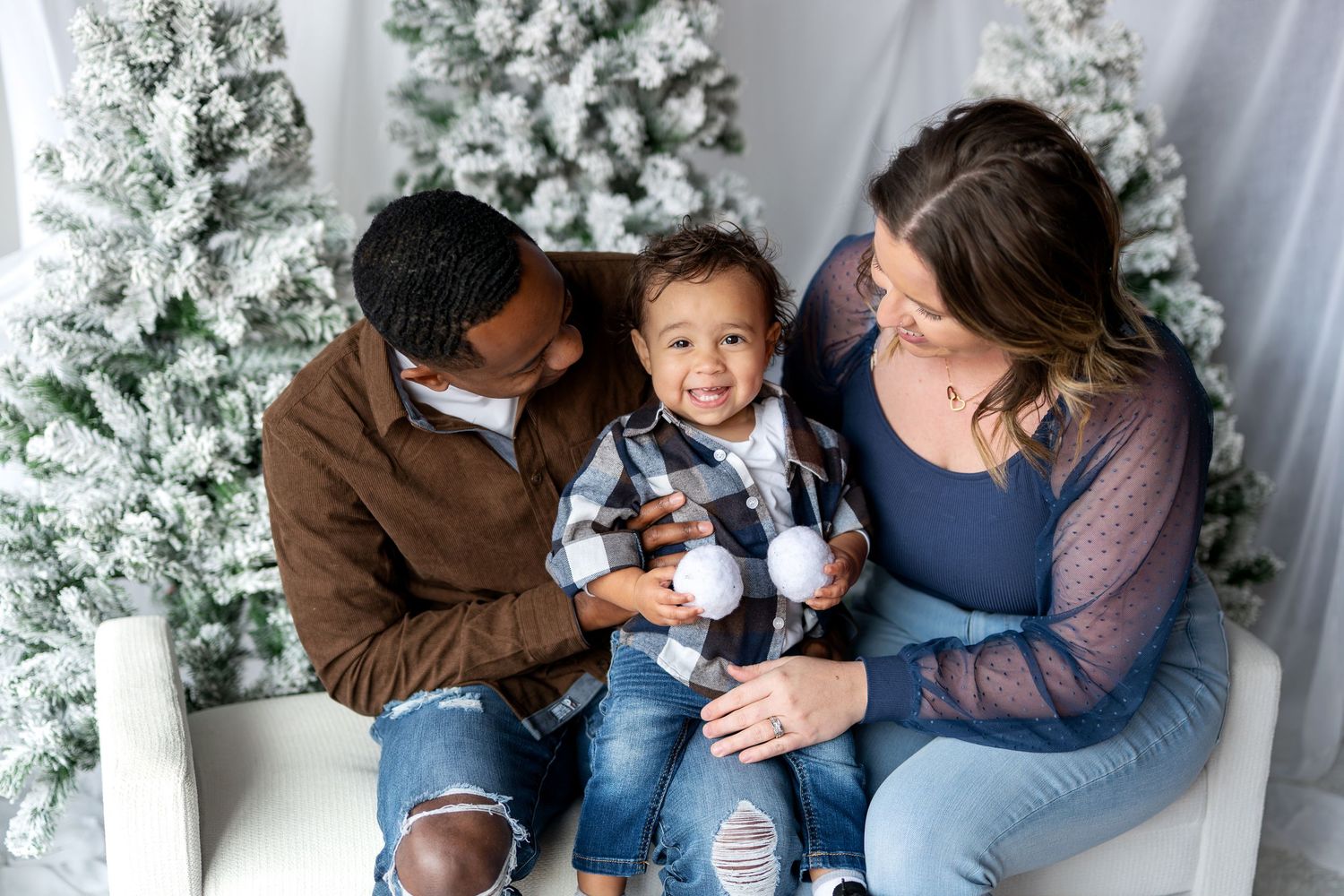 Happy interracial family with toddler son sitting together among snow-flocked Christmas trees.