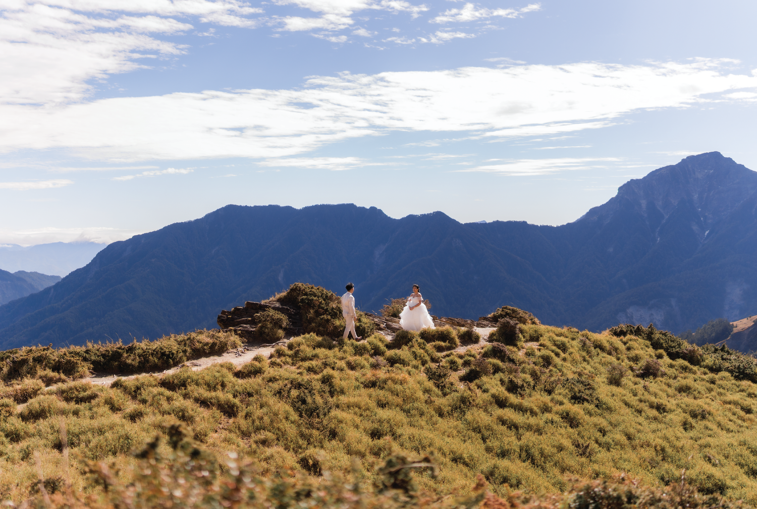 A panoramic mountain landscape with golden grass and rugged peaks under a cloudy blue sky.