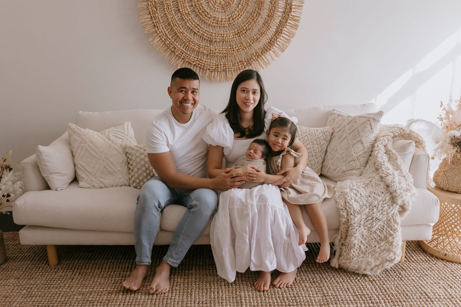A family sits together on a cream-colored couch with decorative pillows and a woven wall hanging above.