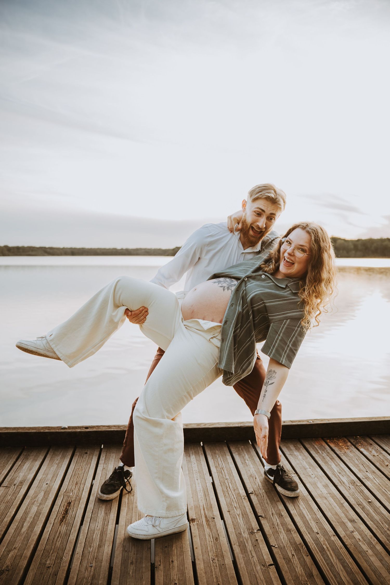 Happy couple playfully posing on a wooden dock by a lake at sunset, woman lifting her leg while man holds her waist.
