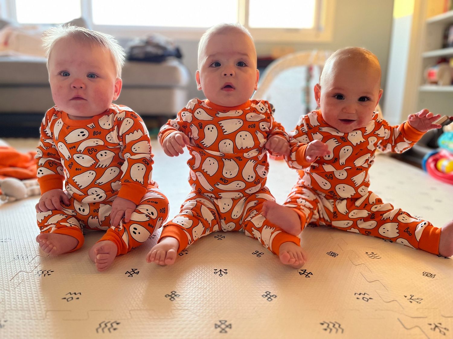 Three baby triplets in matching orange Halloween ghost pajamas sitting together on a white play mat.
