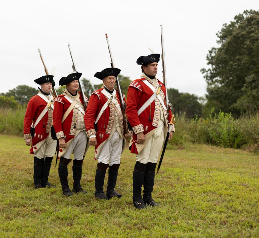 Colonial yorktown reenactment soldiers wearing red and white uniforms in black boots, holding rifles, while lined up