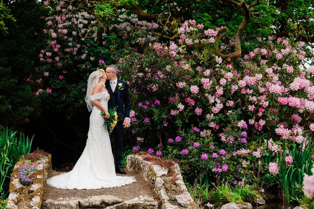 Black and white photo of a bride and groom posing on stone steps surrounded by lush greenery on their wedding day.