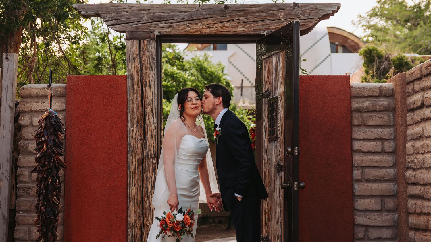 A bride and groom share a kiss under a rustic wooden archway with red adobe walls and lush greenery at their outdoor wedding.