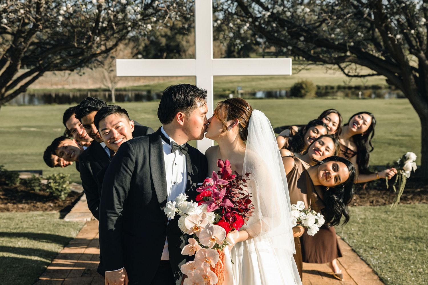 Bride and groom kissing at outdoor wedding ceremony with white cross, surrounded by cheering bridesmaids and groomsmen.