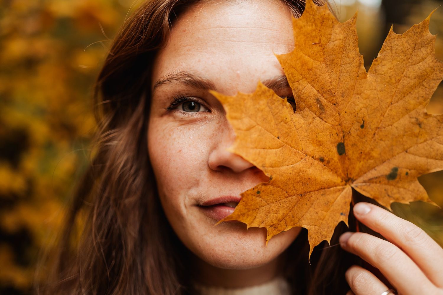 Person holding a large orange autumn maple leaf in front of their face against a blurred fall background.