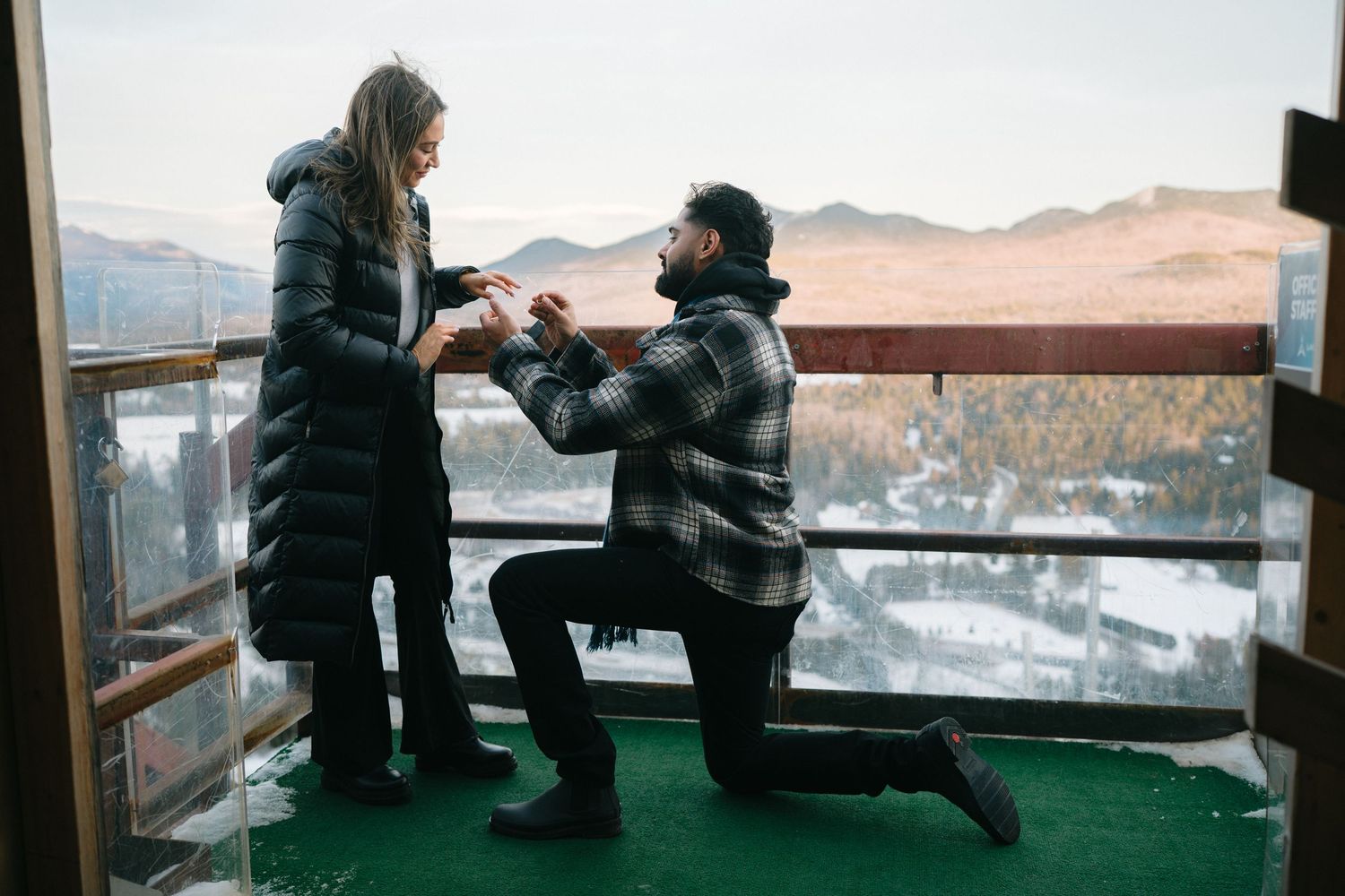 A couple shares a romantic proposal moment on a scenic mountain observation deck at sunset.