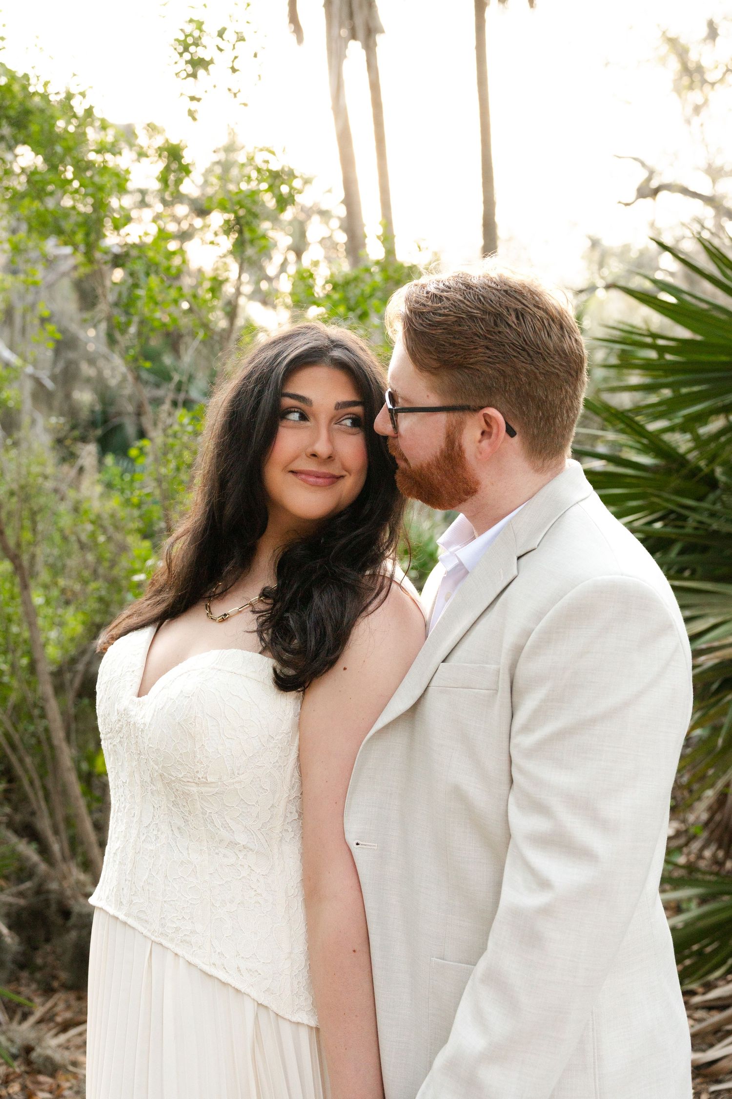 A bride in a white lace gown smiles as her groom in a beige suit whispers in her ear outdoors.