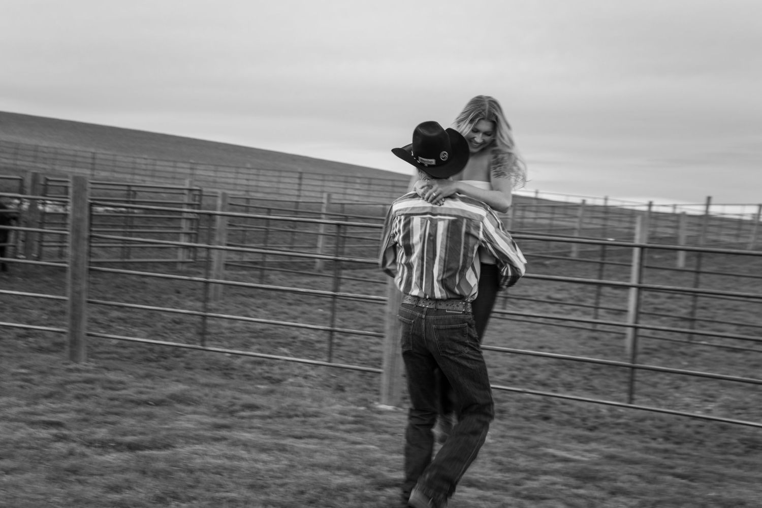 A couple embraces in a black and white photo near a ranch fence, man wearing cowboy hat and striped shirt.