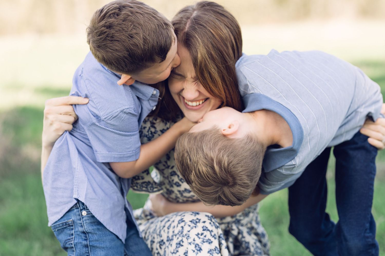 Joyful mother laughing as two young boys embrace her in a sunny green outdoor setting.