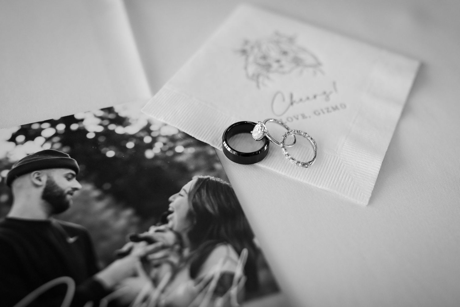Black and white flat lay of wedding rings resting on a personalized cocktail napkin beside a couple's engagement photo.