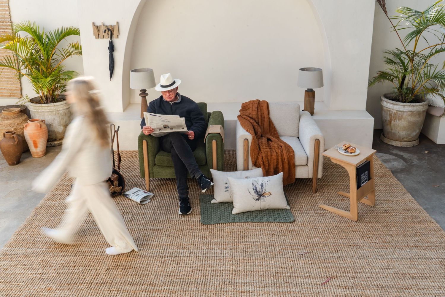 Man in white hat reads newspaper in green chair surrounded by LYBL furniture, cushions, and plants on a jute rug.