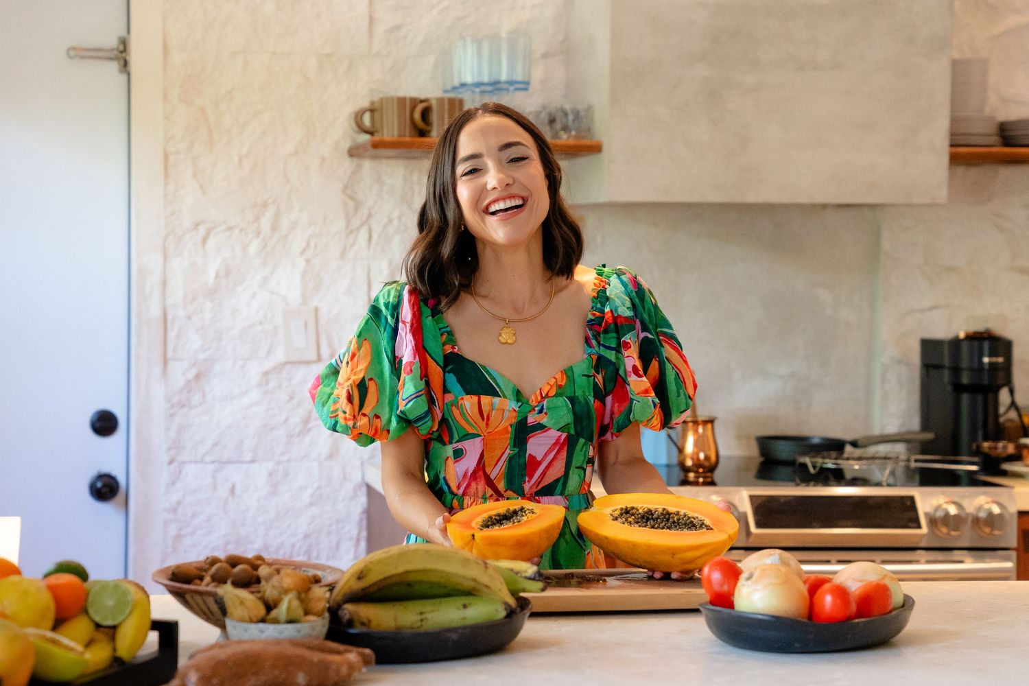 Woman in colorful floral dress smiling in kitchen with fresh tropical fruits including papaya on counter.