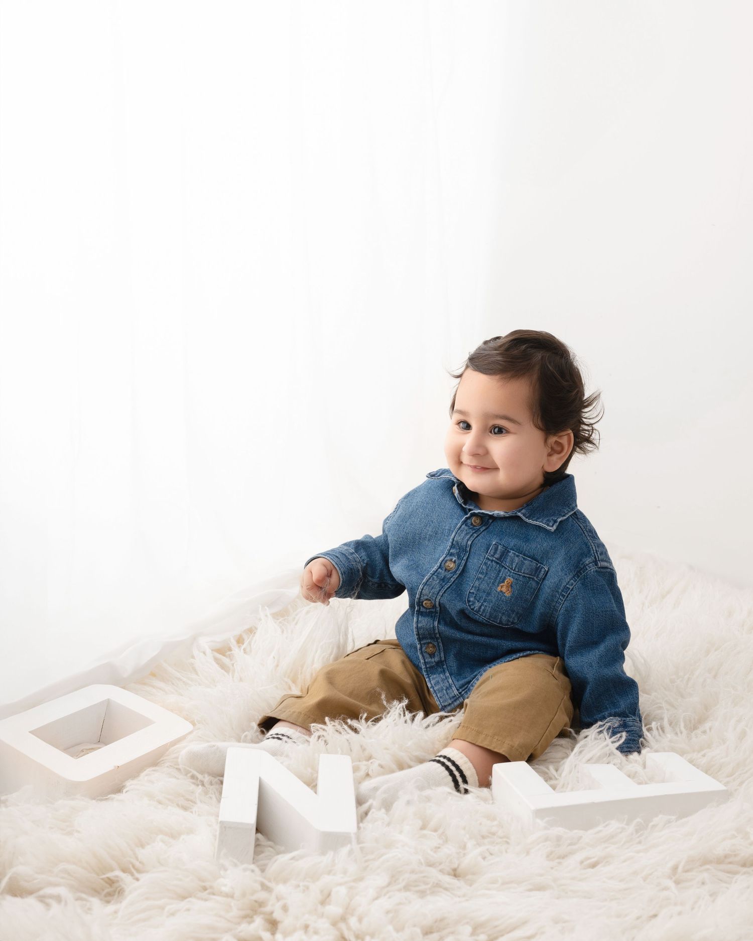 A smiling baby in a denim shirt sits on a fluffy white rug with white letter blocks spelling ONE for a first birthday photo.