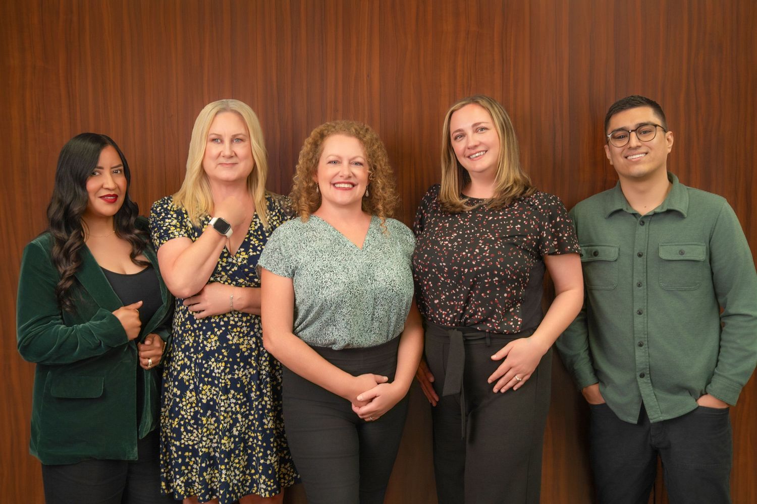 Five professional colleagues smiling together in front of a wooden wall in a modern office setting.