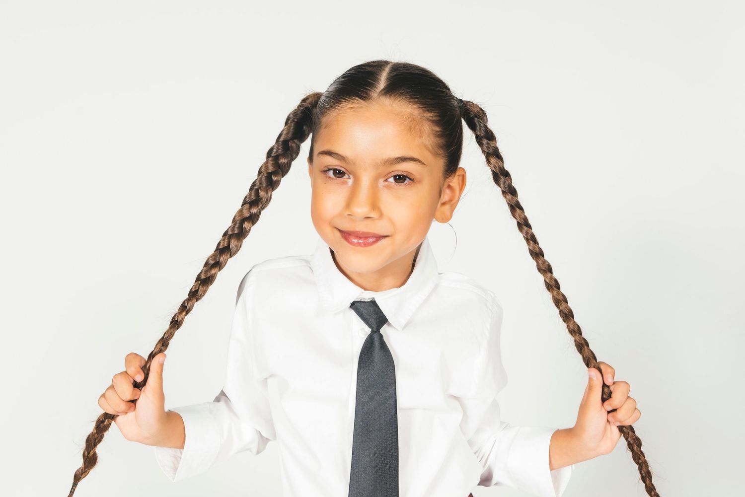 Young girl in white shirt and tie holding long braided pigtails, smiling against white background.