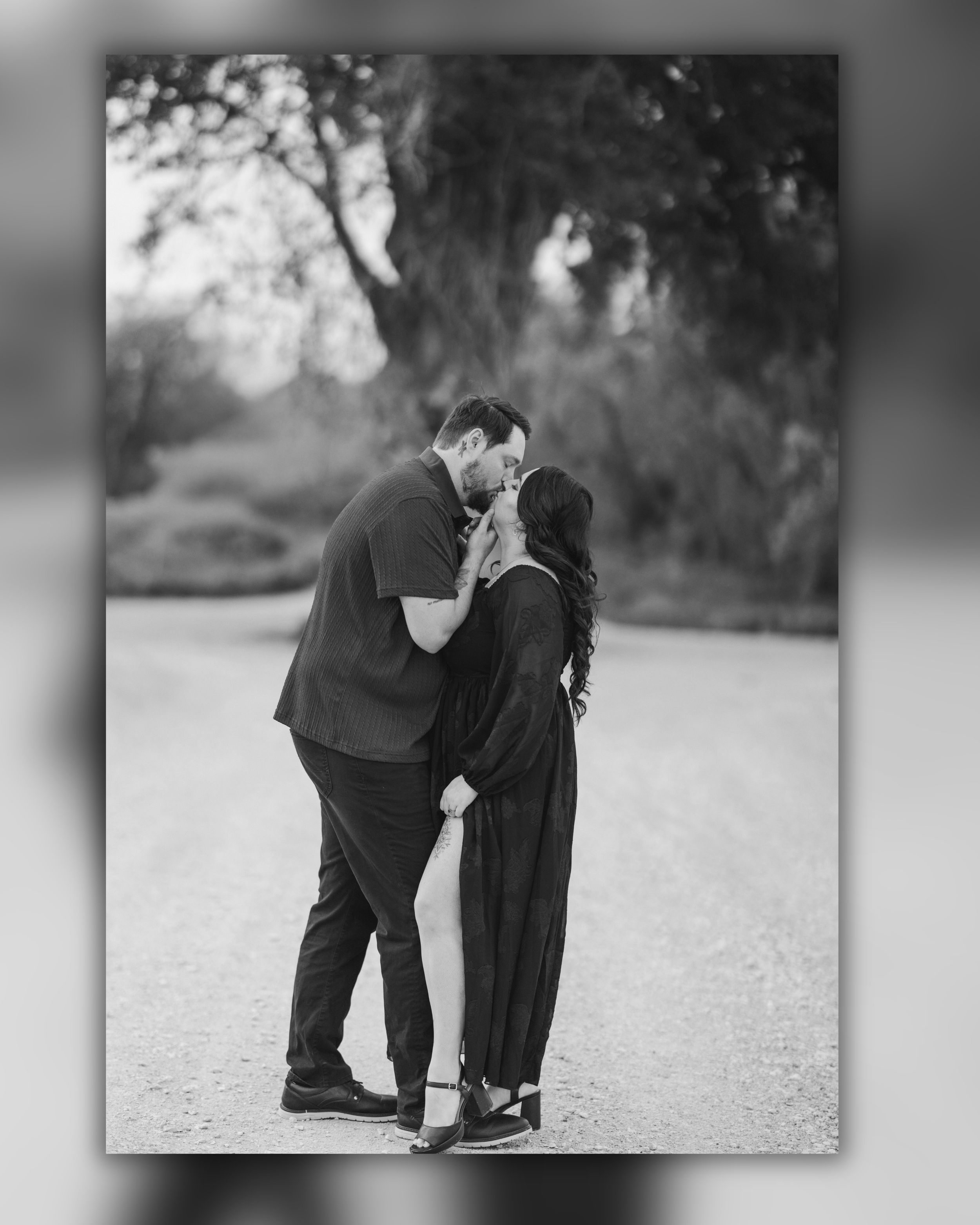 Black and white photo of a couple sharing a romantic kiss outdoors on a gravel path surrounded by lush trees.