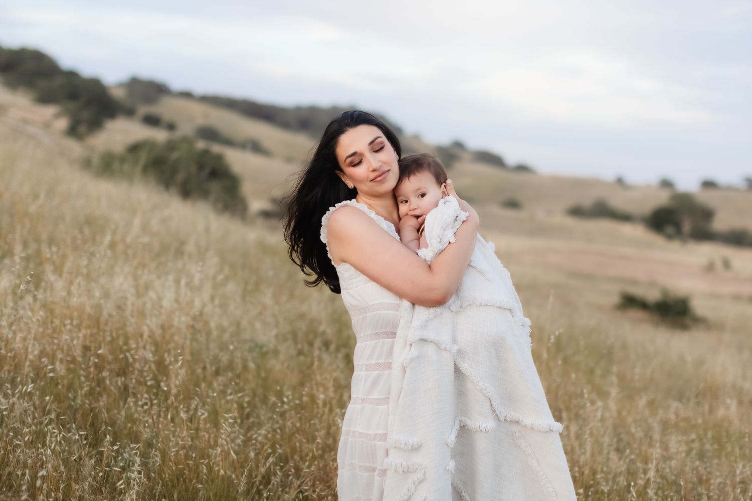 Dark haired mother wears white lace dress snuggles baby in white blanket in grassy field with rolling hills. Concord family photographer