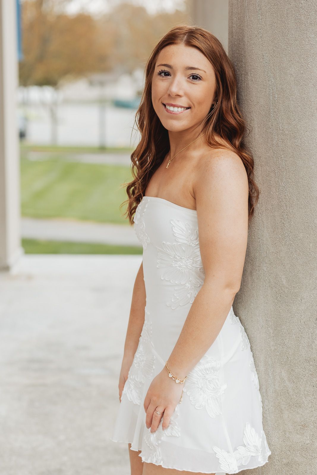 Young woman in white strapless lace mini dress leaning against column, smiling outdoors.