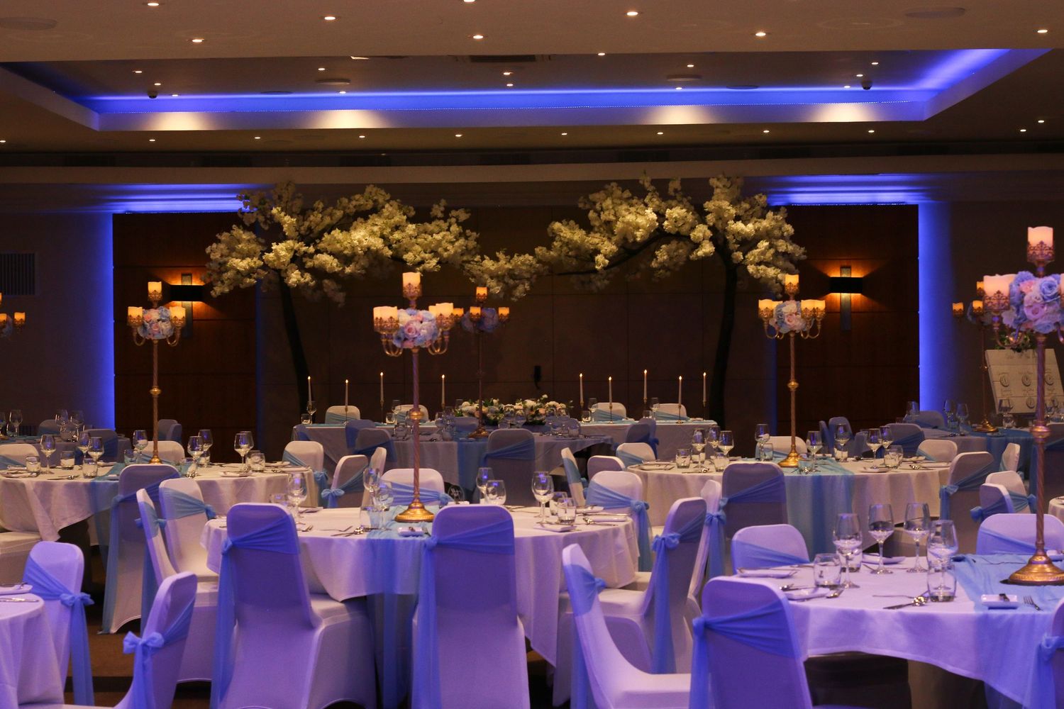 Elegant wedding reception venue with blue uplighting, white chair covers, floral centerpieces, and round tables set for a formal dinner.