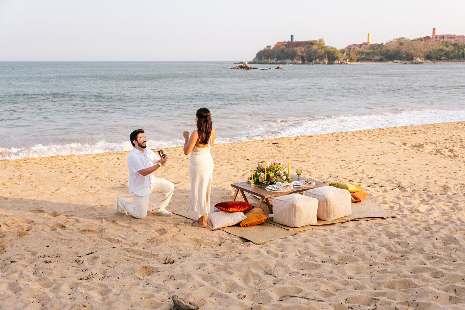 A romantic beach proposal setup with picnic blanket, flowers and decor along the sandy shoreline near coastal cliffs.