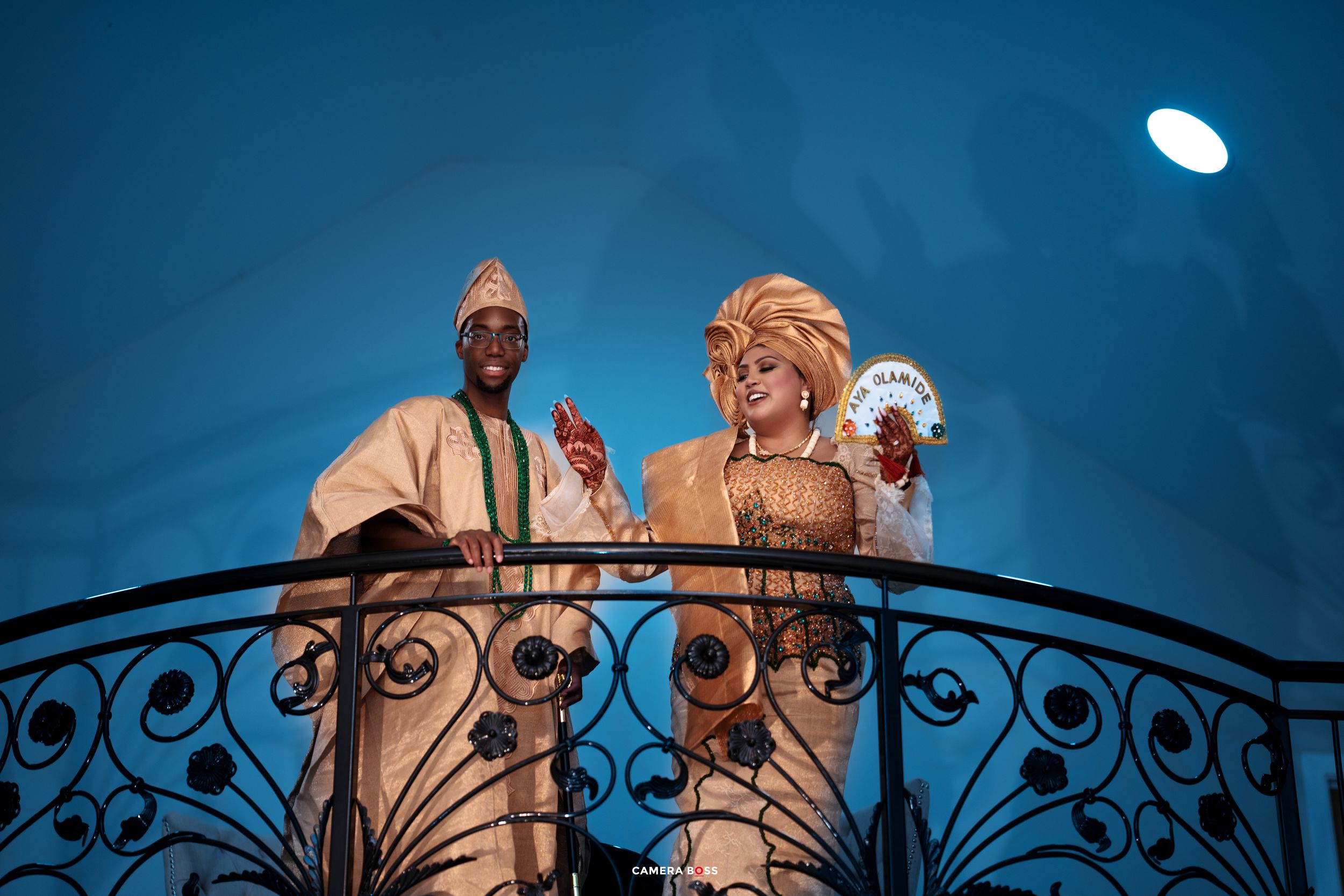 Couple in elegant traditional African attire standing on ornate balcony under blue evening sky.