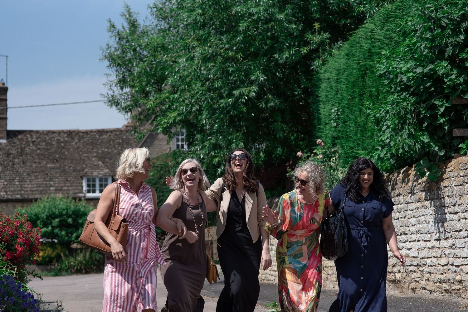 A group of people dance and celebrate together in a garden setting with stone walls and greenery in the background.