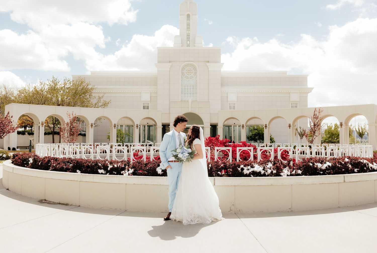 Bride in white gown with veil and groom in light blue suit walking hand in hand through a lush green park.